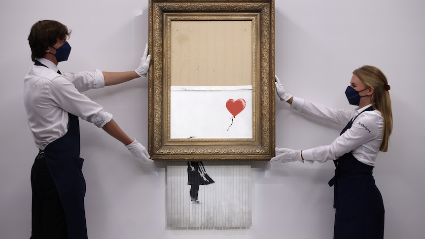 Two people hold a Banksy art on the wall 