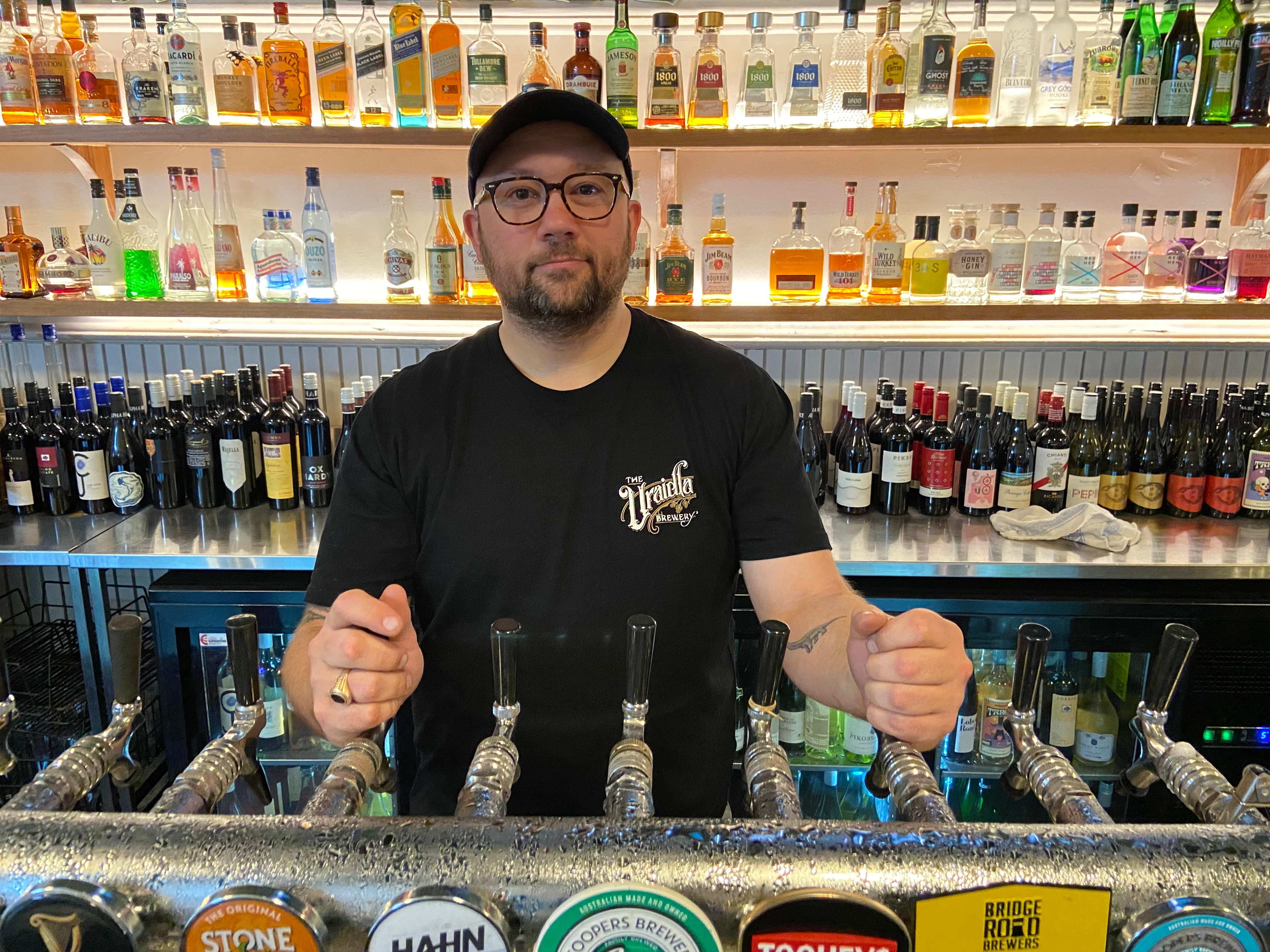 A man in a black t-shirt and cap stands behind a bar with bottles of alcohol all around him