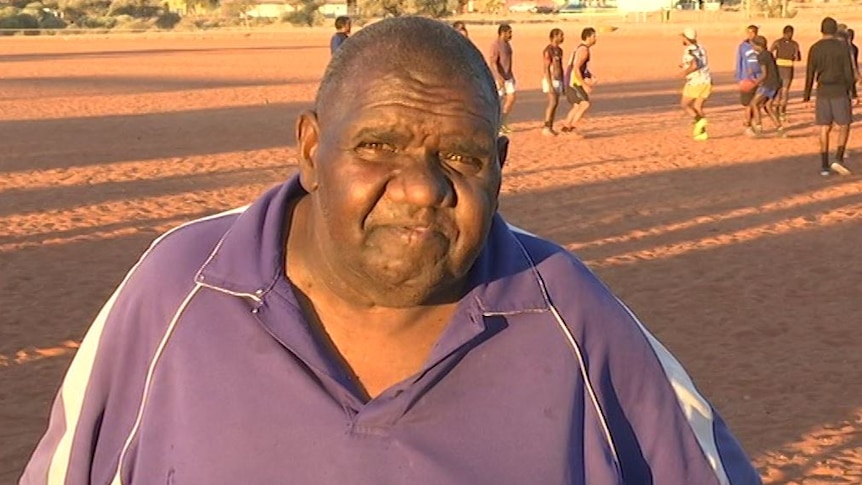 Indigenous man Phillip Alice looks on as the players train behind him on a dirt football oval.