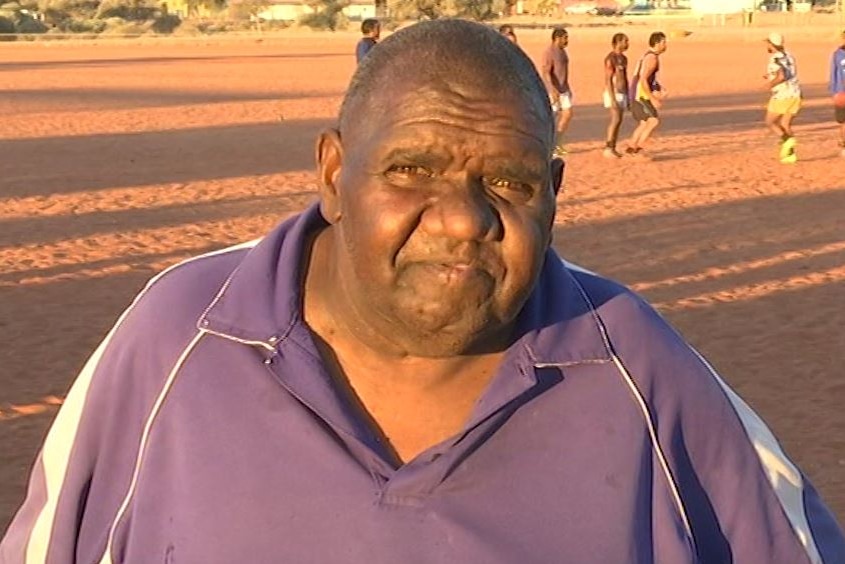 Man looks on as the players train behind him on a dirt football oval.