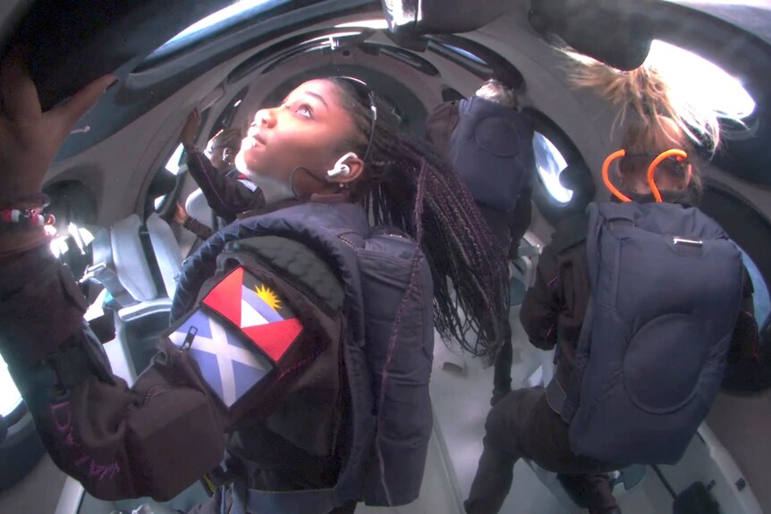 A woman wearing a black space suit looking out a a circle window while floating in a spacecraft tube