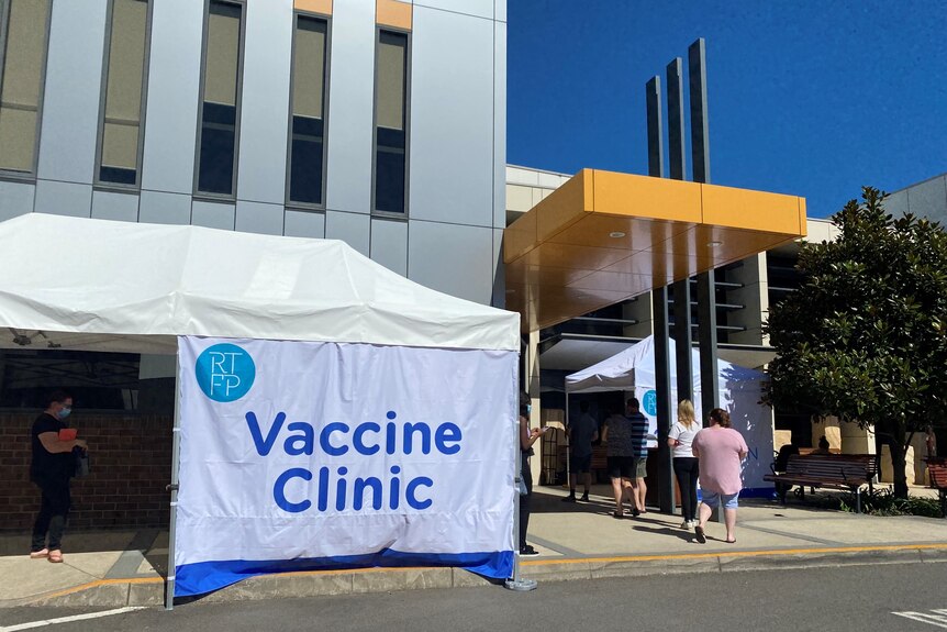 A sign reading "vaccination clinic" outside a large building, with a line people waiting outside.