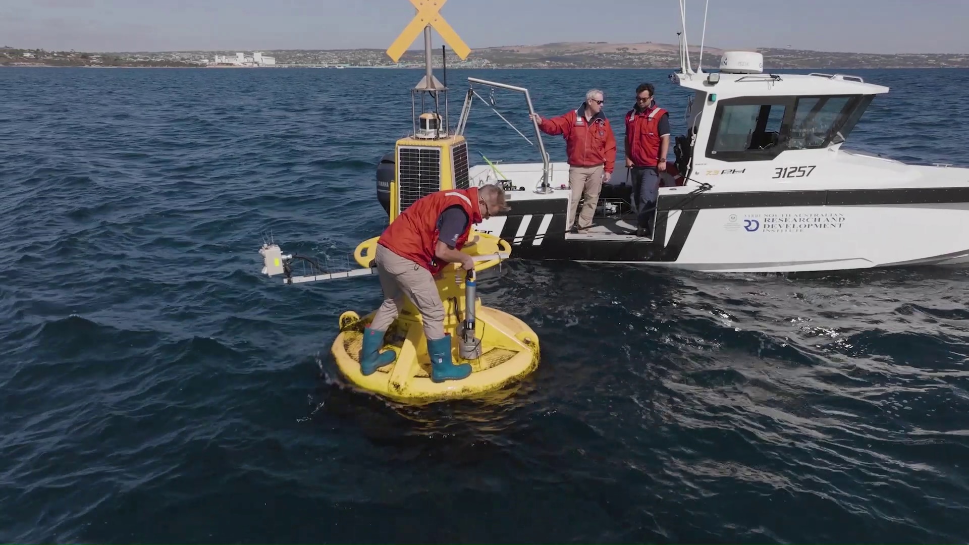 two men stand on a boat in the ocean and watch an other man standing on a yellow buoy