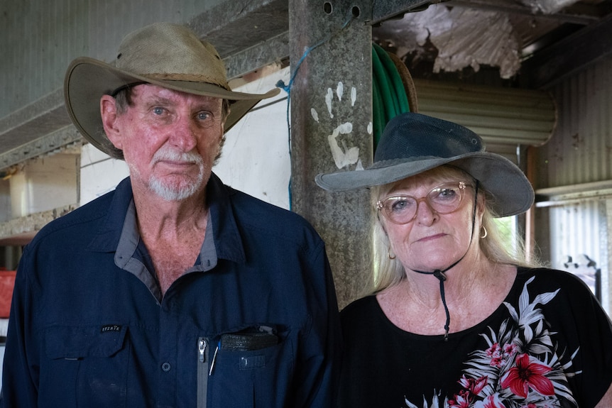 Gary and Sandra (left to right stand in their shed and look into the camera. Both wear hats.