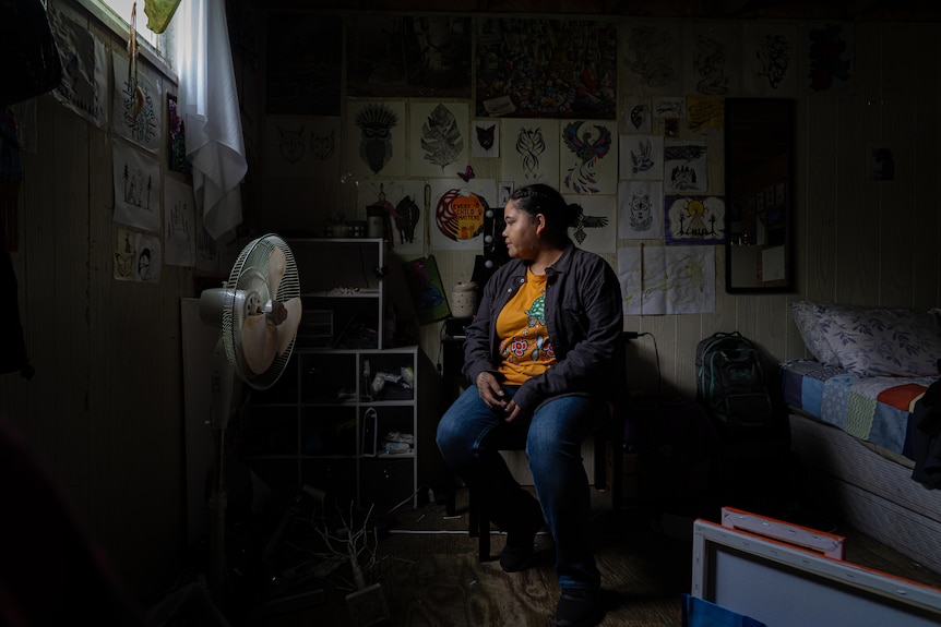 A young First Nations Canadian woman sits in her dark bedroom. Art adorns the walls.