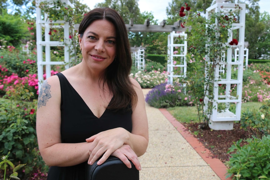 Rachael smiles into the camera, standing in the rose garden. She wears a black dress and has a tattoo.