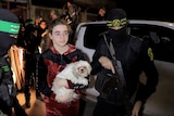 A young person holding a small dog and standing outside between two armed men wearing balaclavas, standing near cars.
