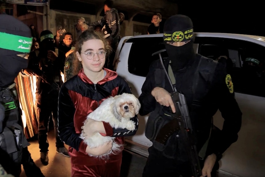 A young person holding a small dog and standing outside between two armed men wearing balaclavas, standing near cars.