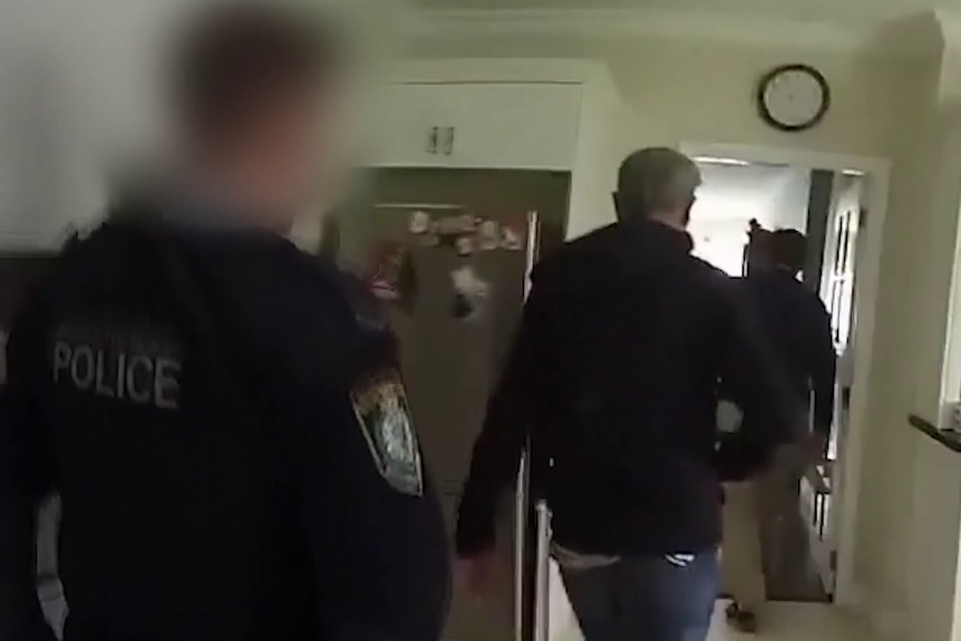 Man walking through a kitchen with police officer behind him.