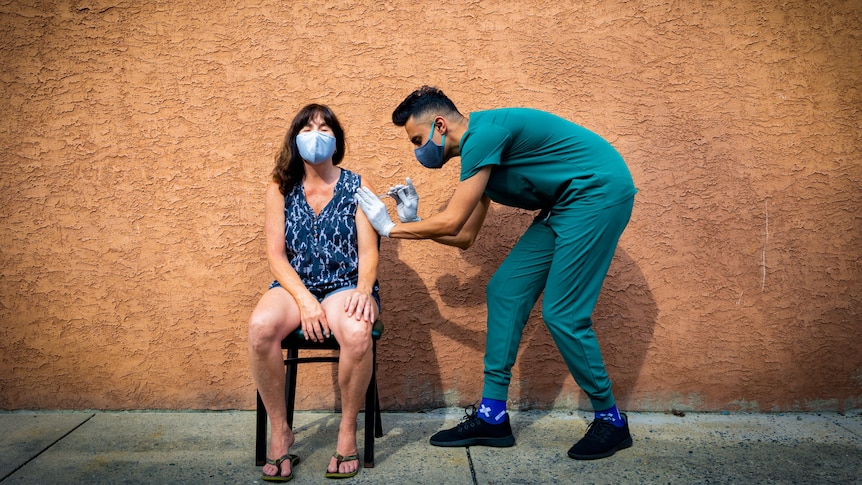 A woman in a blue mask sits in a chair against a terracotta wall while a health worker in scrubs injects a needle into her arm