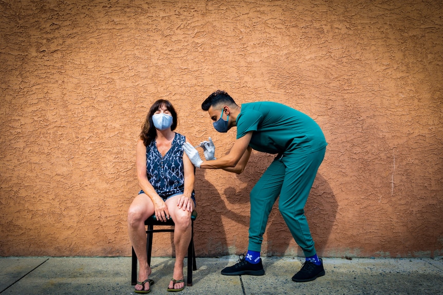 A woman in a blue mask sits in a chair against a terracotta wall while a health worker in scrubs injects a needle into her arm