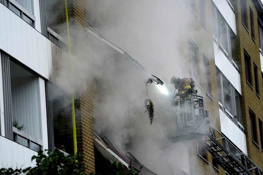 A firefighter stands on the ladder of a fire truck as smoke billows from an apartment building.