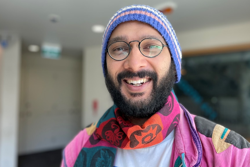 A smiling man wearing a pink jacket, scarf and blue striped beanie.