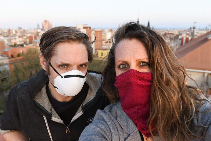 Andrew Burden and Ali Cameron, with their nose and mouths covered with masks, stand on rooftop of Barcelona house.
