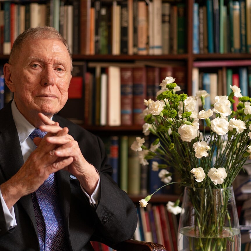 Michael Kirby sitting withbooks on shelves behind him and a bouquet of flowers next to him.