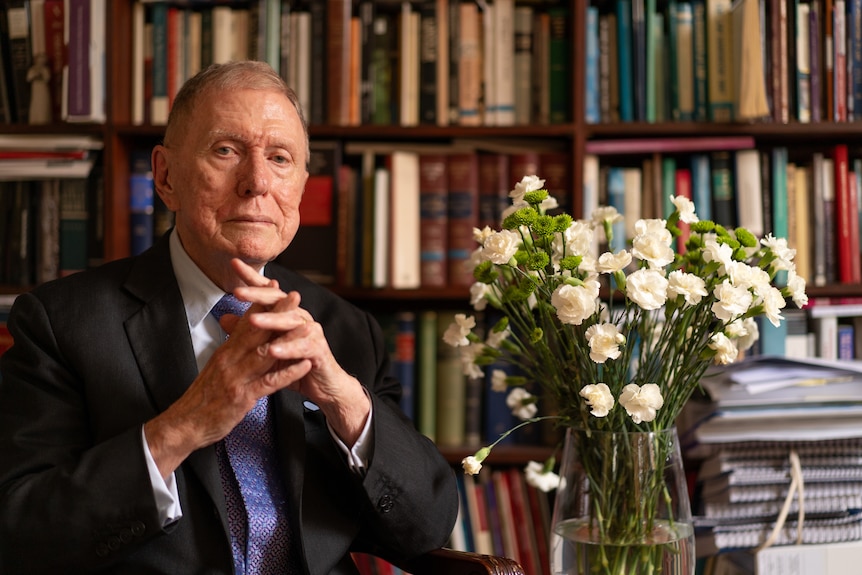 Michael Kirby sitting withbooks on shelves behind him and a bouquet of flowers next to him.
