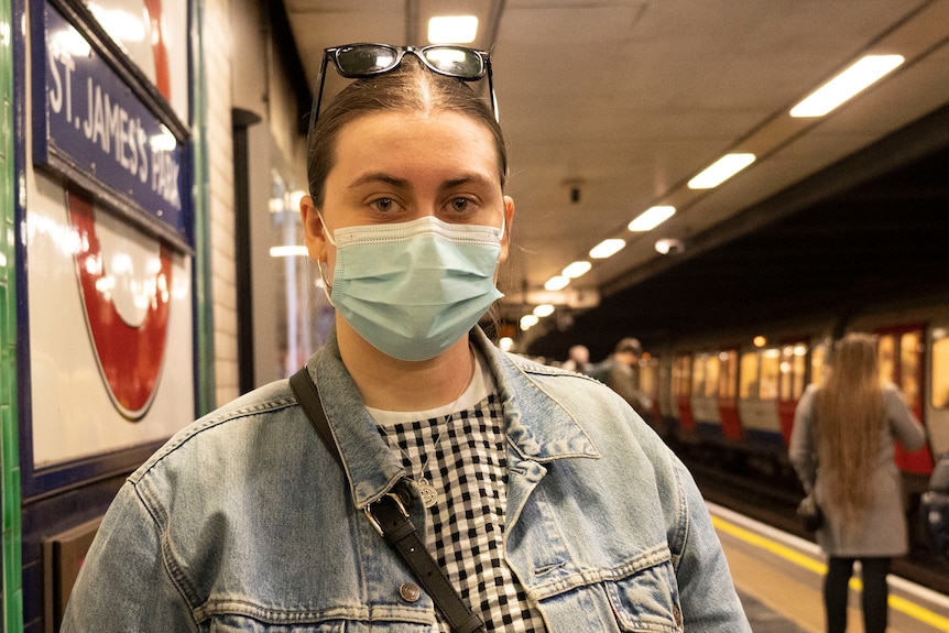 A woman wearing a denim jacket and checkered top wears a mask and sunnies on her head.