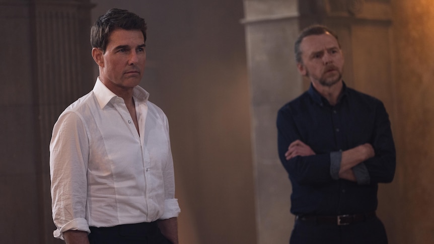 Tom Cruise, a white brunette man in a white shirt, and Simon Pegg, a white blonde man in navy shirt, look quizzically off camera