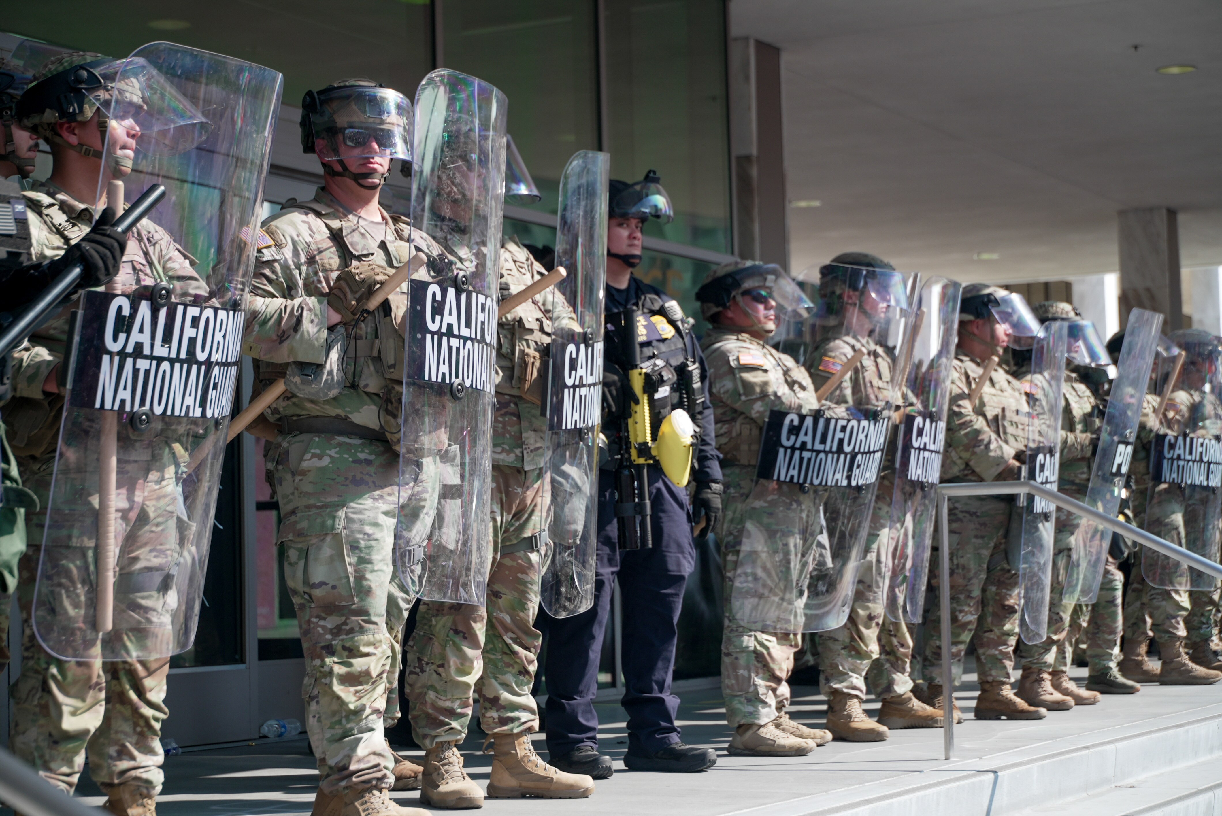 A row of men in military wear hold transparent shields that say 'California National Guard'.