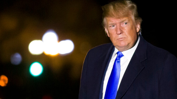 US President wearing a dark blue suit and bright blue shiny tie looking sideways with helicopter lights in the background