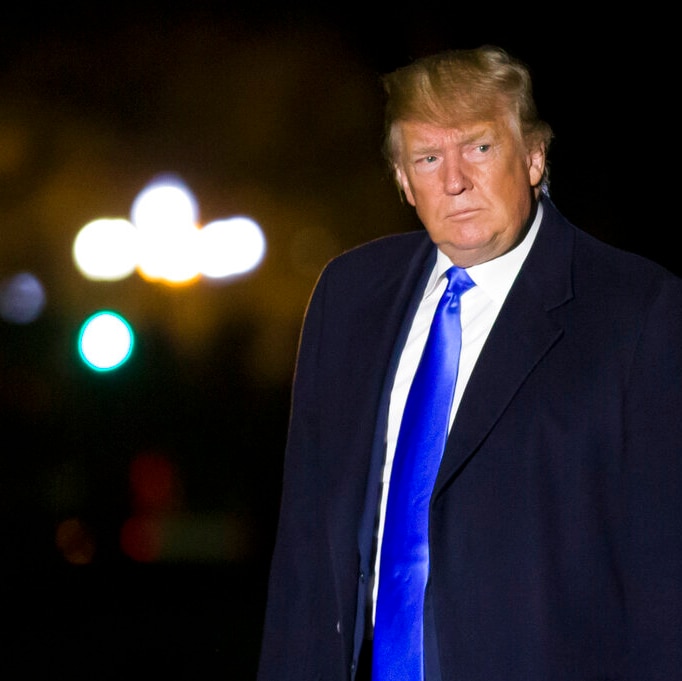 US President wearing a dark blue suit and bright blue shiny tie looking sideways with helicopter lights in the background