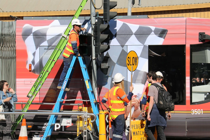 A man stands on a ladder at a tram stop as he works on the pedestrian traffic lights.