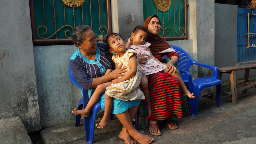 Four-year-old twins Fani and Fina in Perumnas Klender. They are sitting outside on the laps of their mother and another woman.