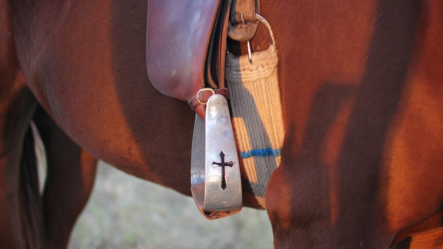 Close-up of a stirrup hanging at a horse's side from the stirrup leather. The stirrup has a cross detailing cut into it.