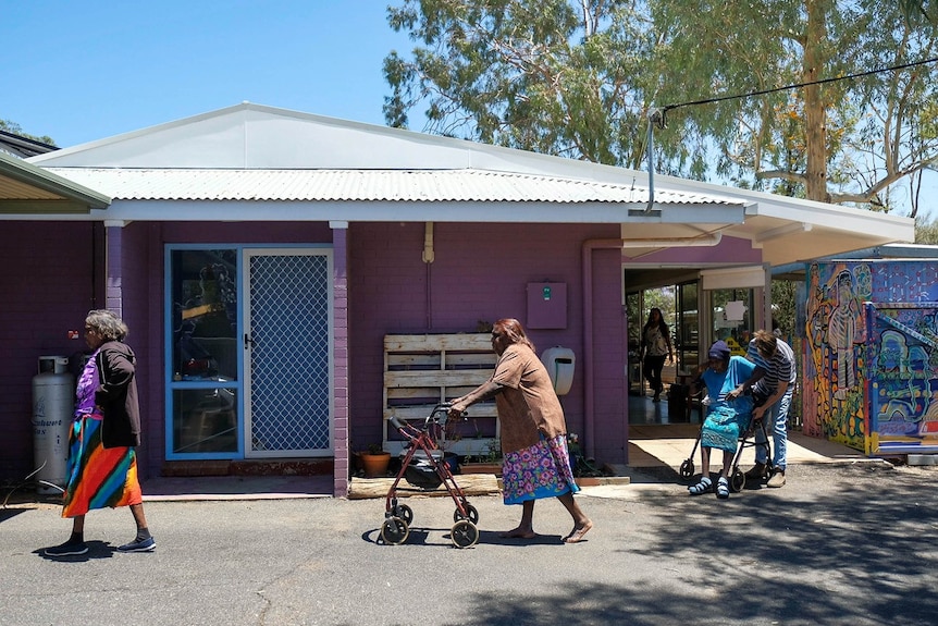Three Indigenous women, two with walkers, walk past the exterior of a purple house