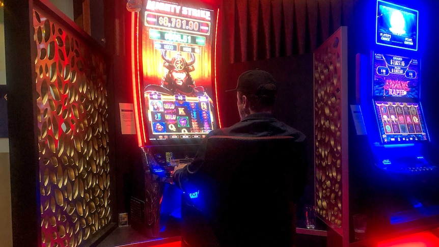Man wearing black shirt and cap sits backed turned to camera at neon pokie machine, with other pokie machine visible