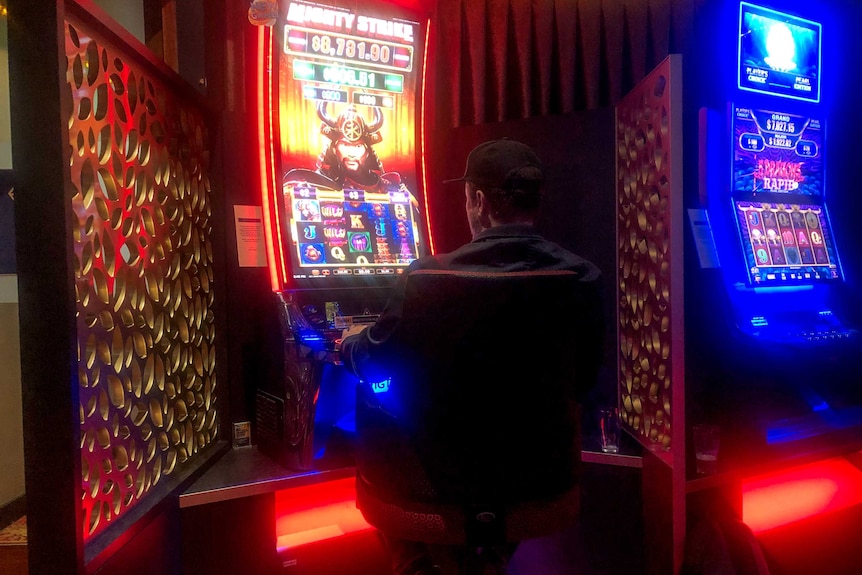 Man wearing black shirt and cap sits backed turned to camera at neon pokie machine, with other pokie machine visible