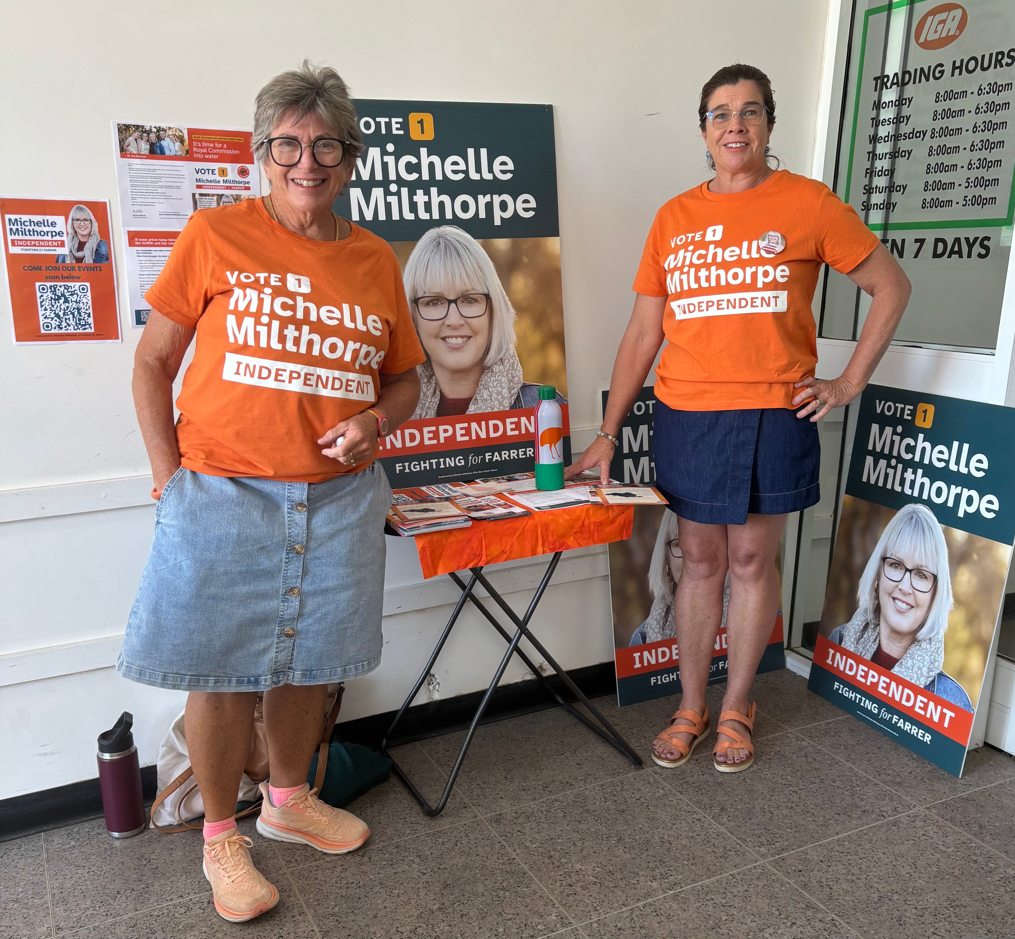 Two women stand together wearing orange t-shirts with white writing, reading 'VOTE 1 Michelle Milthorpe Independent'.