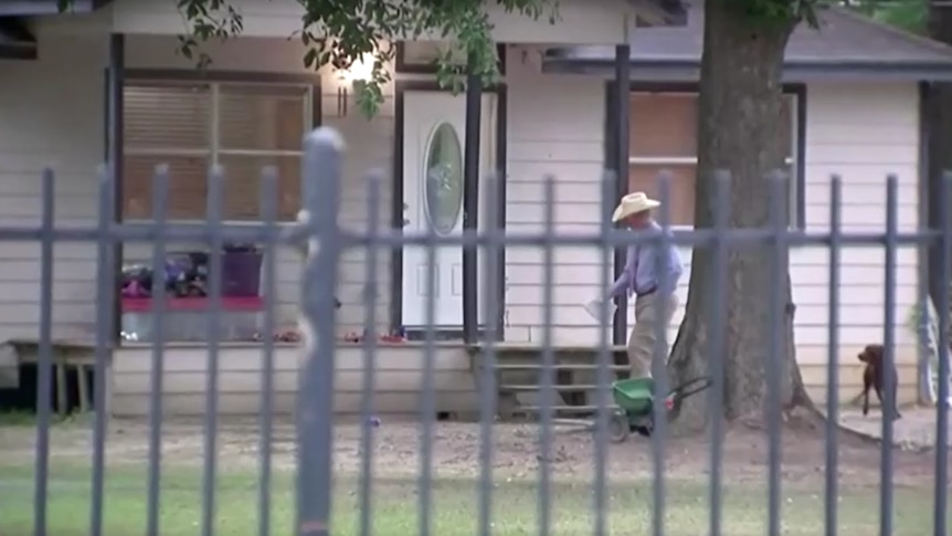 An investigator in a blue shirt and white cowboy hat walks past the entrance to a home behind a barred fence.