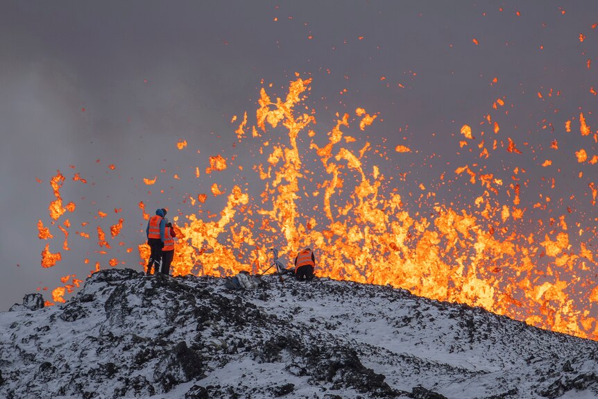 Scientists in orange hi-vis stand in front of lava explosion 