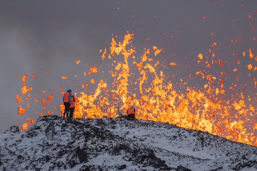 Scientists in orange hi-vis stand in front of lava explosion 