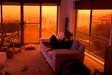 A woman sits on a lounge chair in a smoky room with eerie red sky outside.
