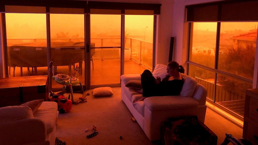 A woman sits on a lounge chair in a smoky room with eerie red sky outside.