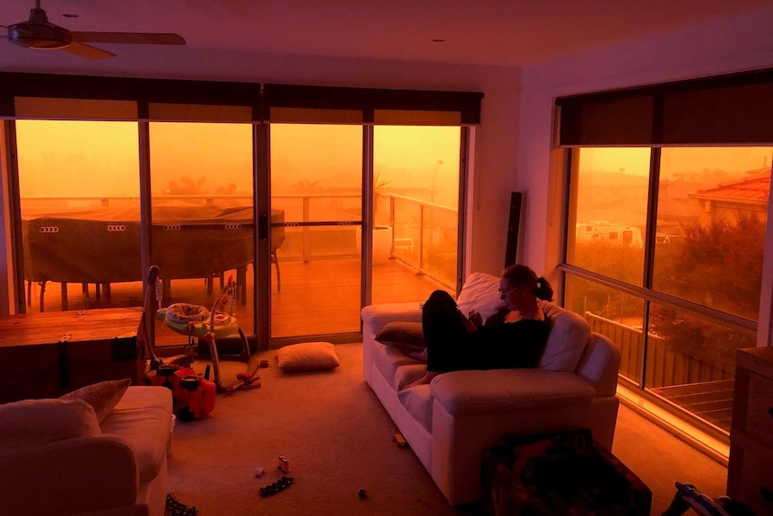 A woman sits on a lounge chair in a smoky room with eerie red sky outside.
