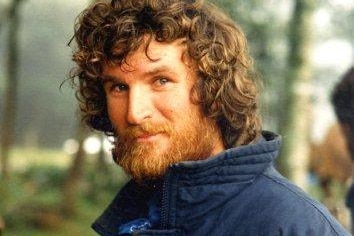 Colour photo of red-headed and bearded man smiling with head turned to side, and blurred greenery in background.