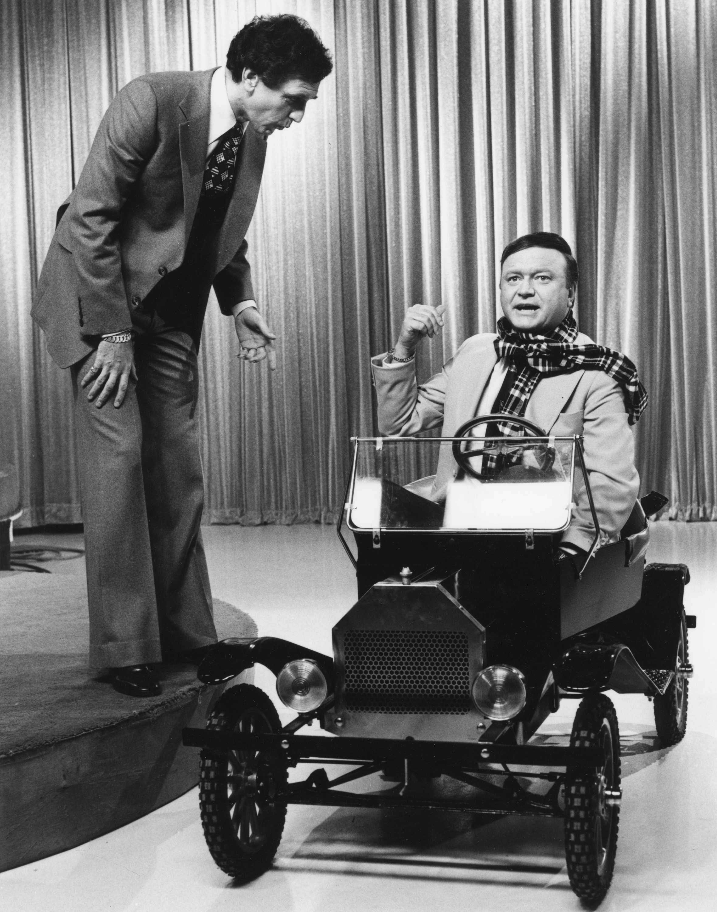 A black and white photo of Don Lane looking down at Bert Newton, who is driving a mini Ford car in a TV studio.