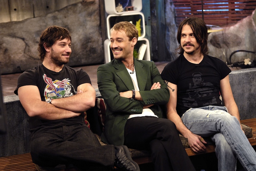 Three members of Silverchair sit on an interview couch. Daniel smiles looking at Chris 