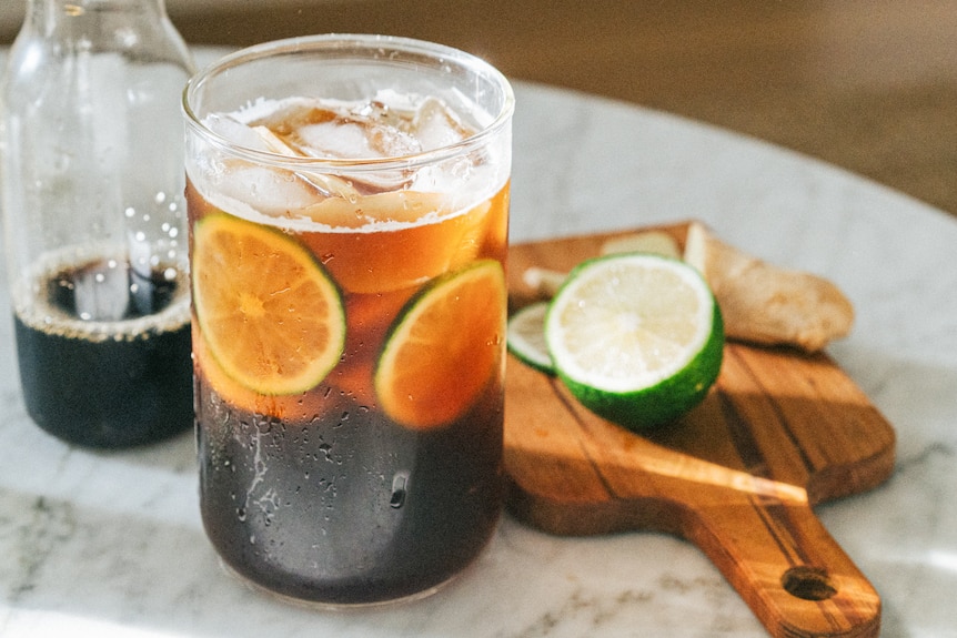 A glass of dark, ice coffee with lime slices inside. It sits on a marble table with a bottle of coffee behind it.