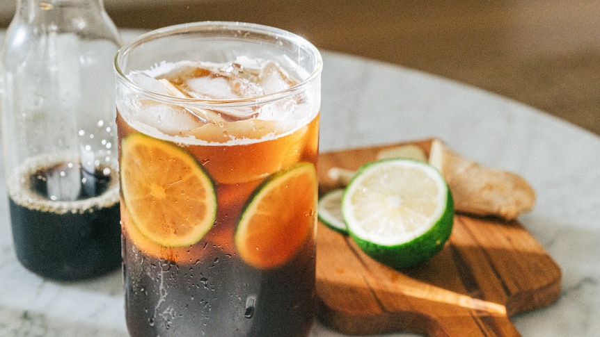 A glass of dark, ice coffee with lime slices inside. It sits on a marble table with a bottle of coffee behind it. 