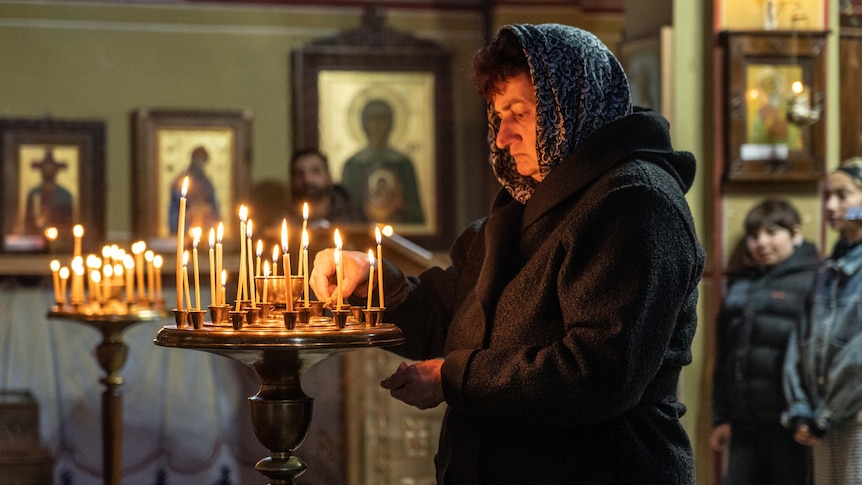 An older woman wearing a headscarf lights a candle inside a church