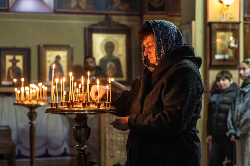 An older woman wearing a headscarf lights a candle inside a church
