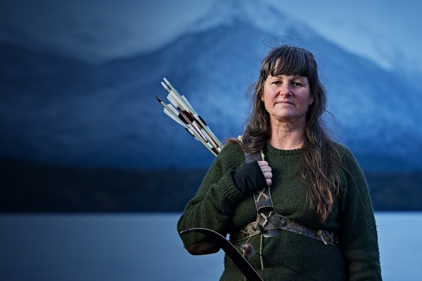 Head and shoulders image of a woman in outdoors cold-weather gear holding a sheaf of arrows in front of a mountain backdrop.