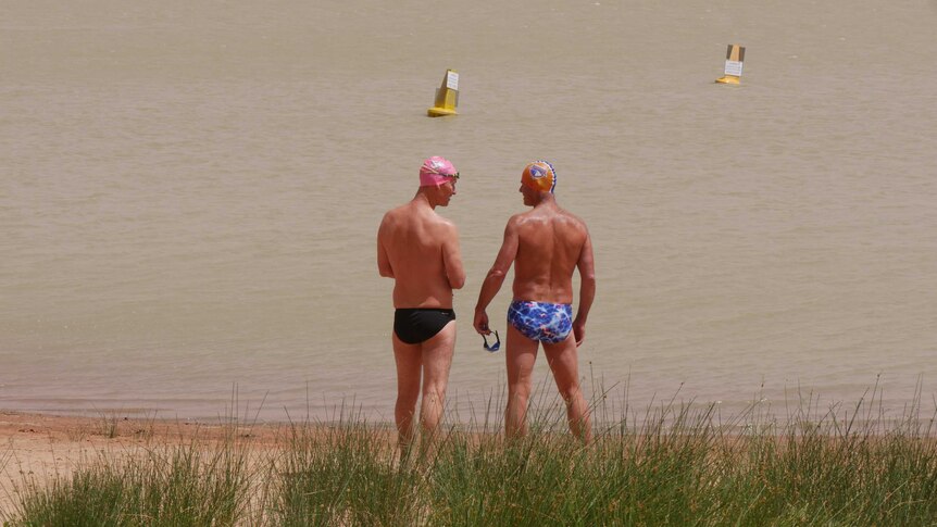 Two men in speedos stand in front of a body of water.