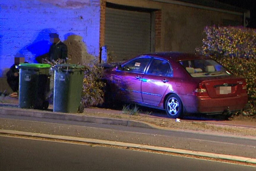 A red Toyota Corolla is seen on a curb between two hedges. Two people lean against a wall behind two green bins