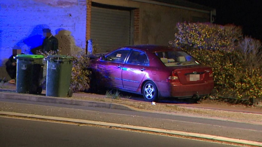 A red Toyota Corolla is seen on a curb between two hedges. Two people lean against a wall behind two green bins