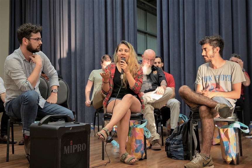 A woman speaks into a mic on a stage surrounded by other people